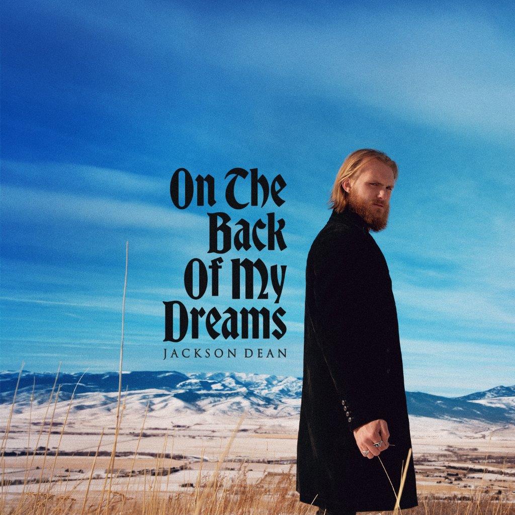 Jackson Dean stands in a field against a blue sky and mountain landscape.