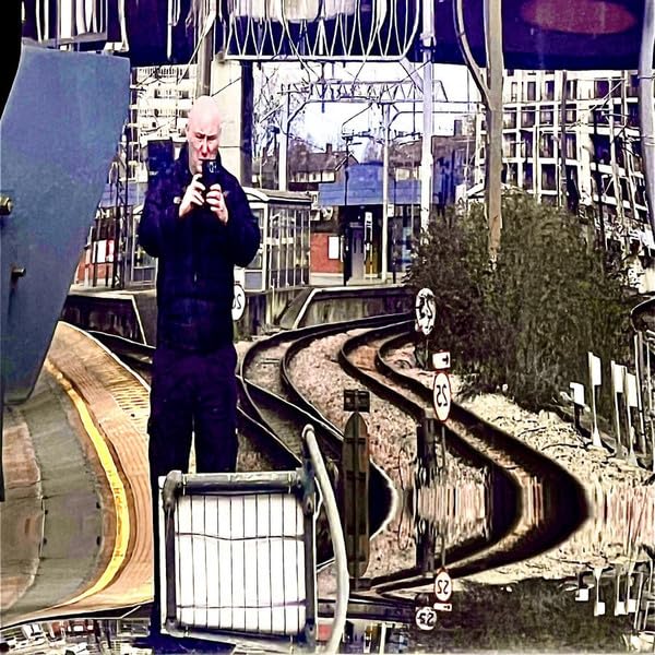 Bald man with a harmonica stands on a railway platform.