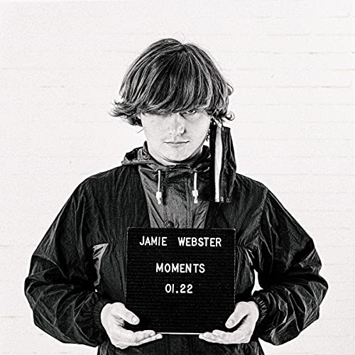 Black and white portrait of Jamie Webster holding a sign reading "Moments.