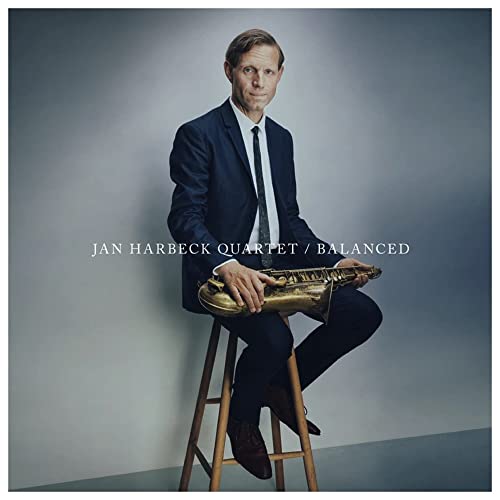 A man in a suit sits on a stool holding a saxophone against grey background.