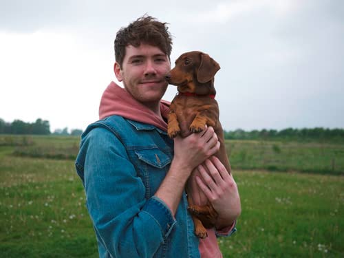 A man in a denim jacket holding a brown dachshund in a grassy field.