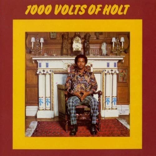 John Holt sits in a chair before a fireplace on this album cover.