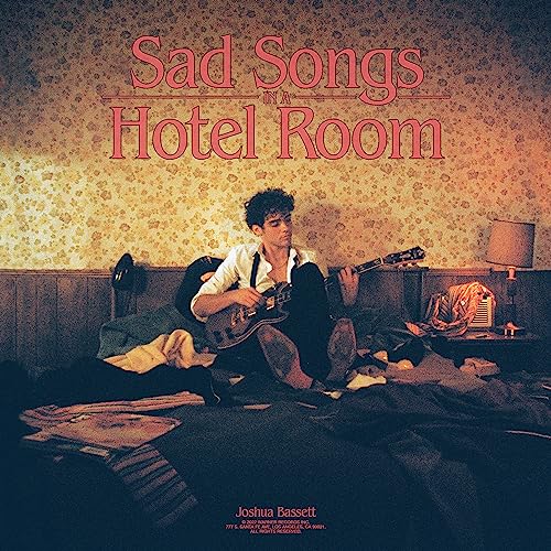 Joshua Bassett sits on a bed playing guitar in a floral-wallpapered hotel room.