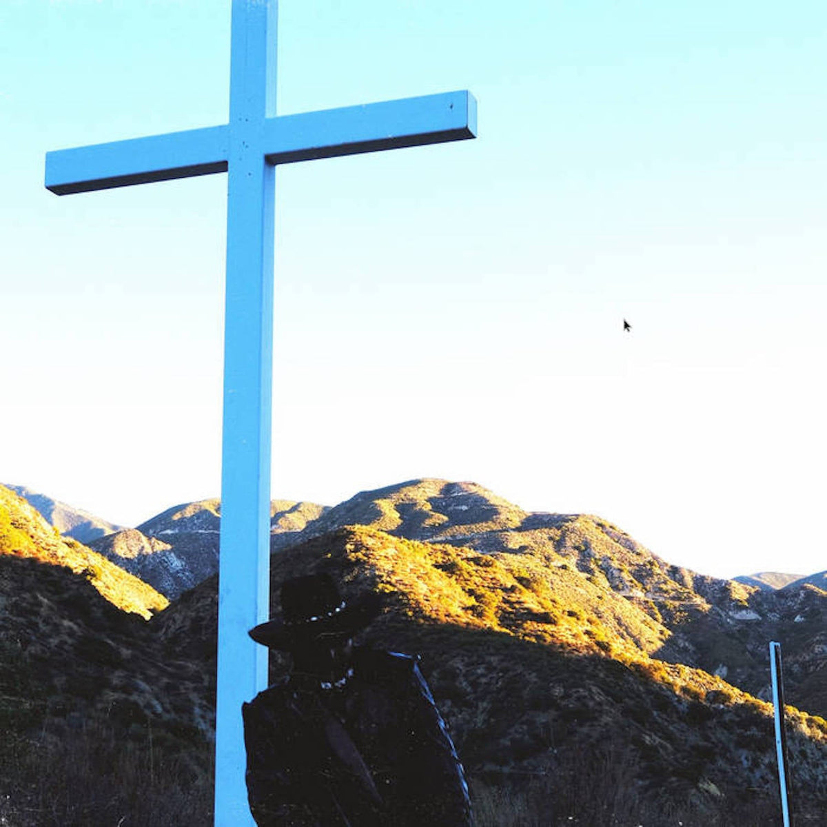 A person in a hat stands before a large white cross against mountain hills.