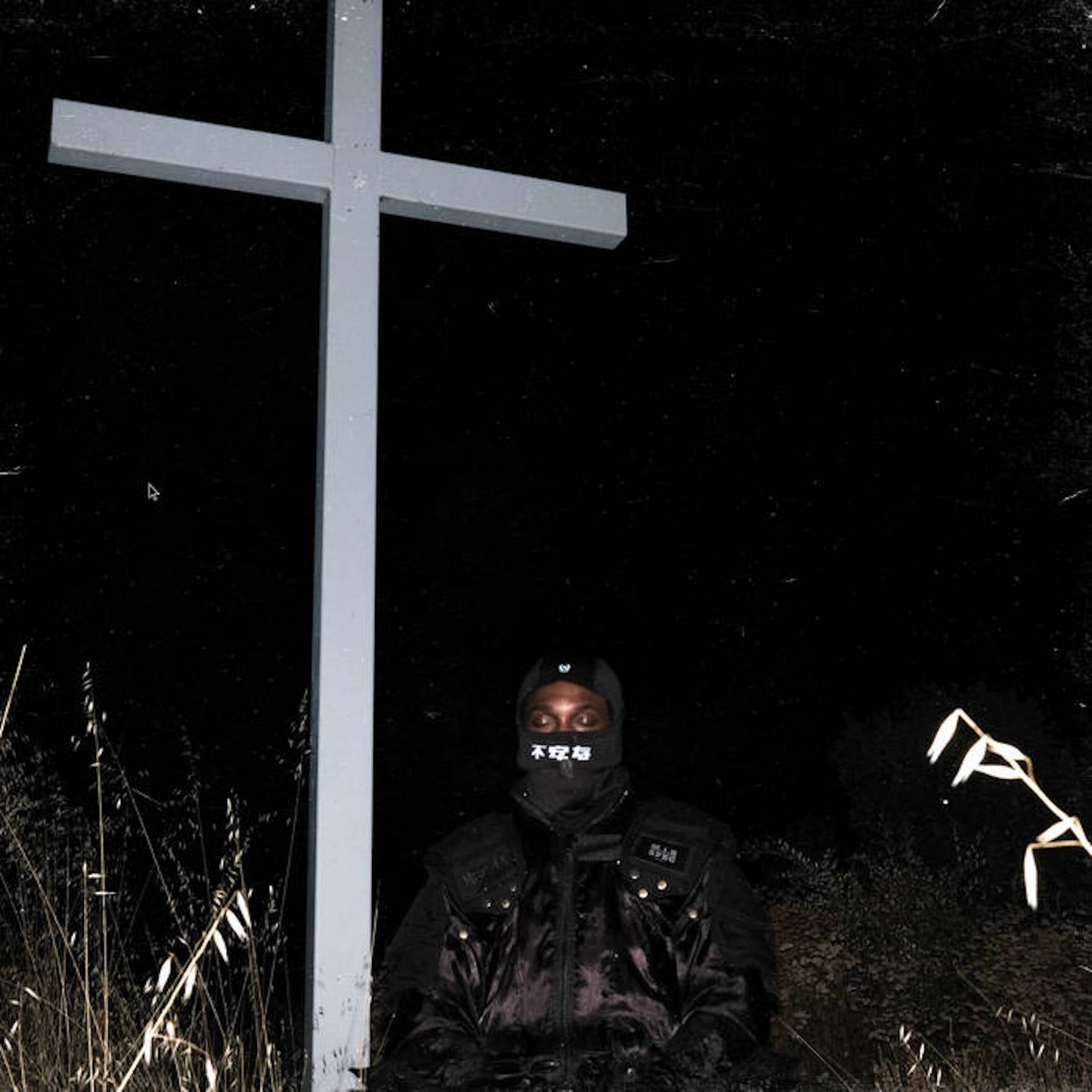 Figure in ski mask stands before a large, weathered wooden cross.