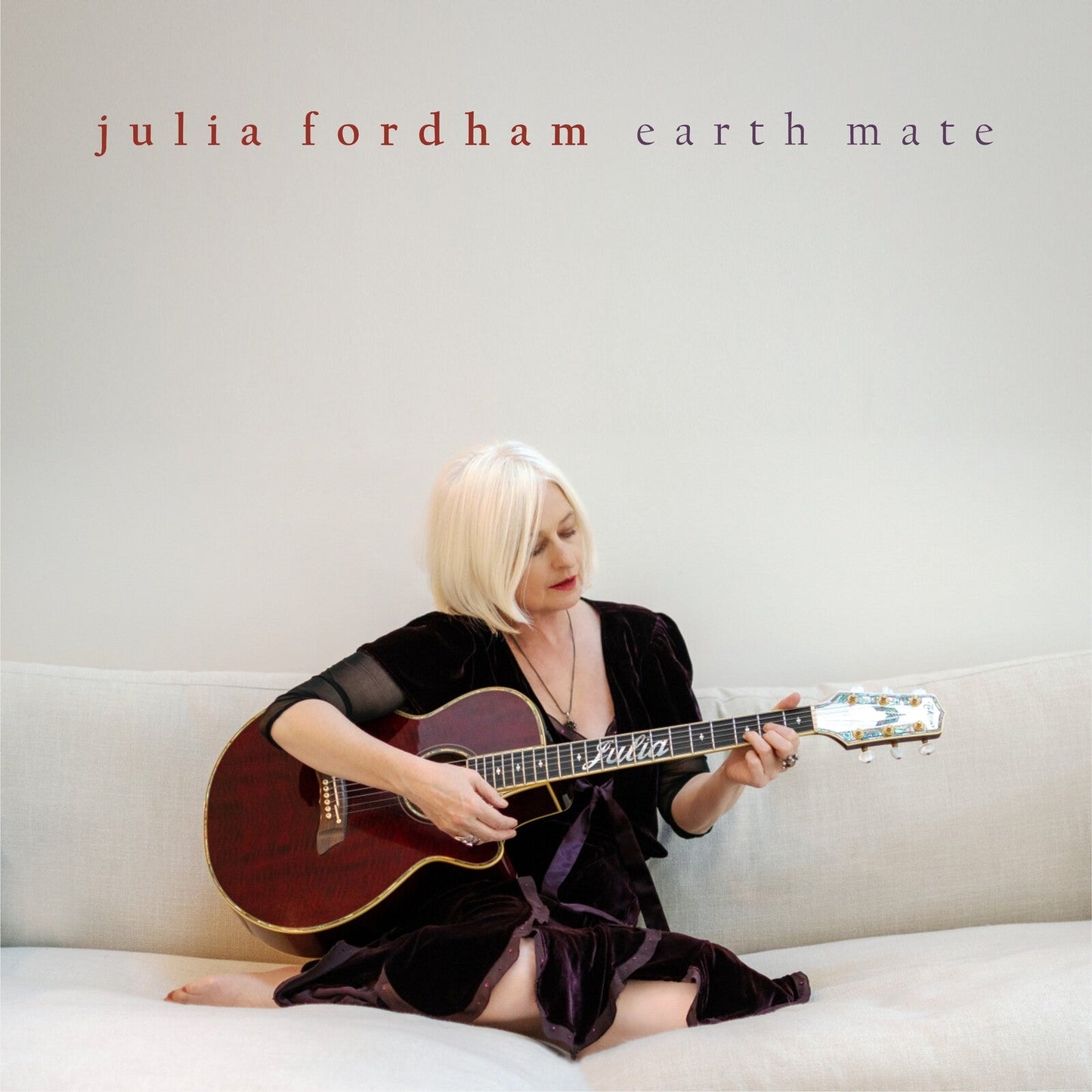 Julia Fordham with blonde hair sits on a sofa playing an acoustic guitar.