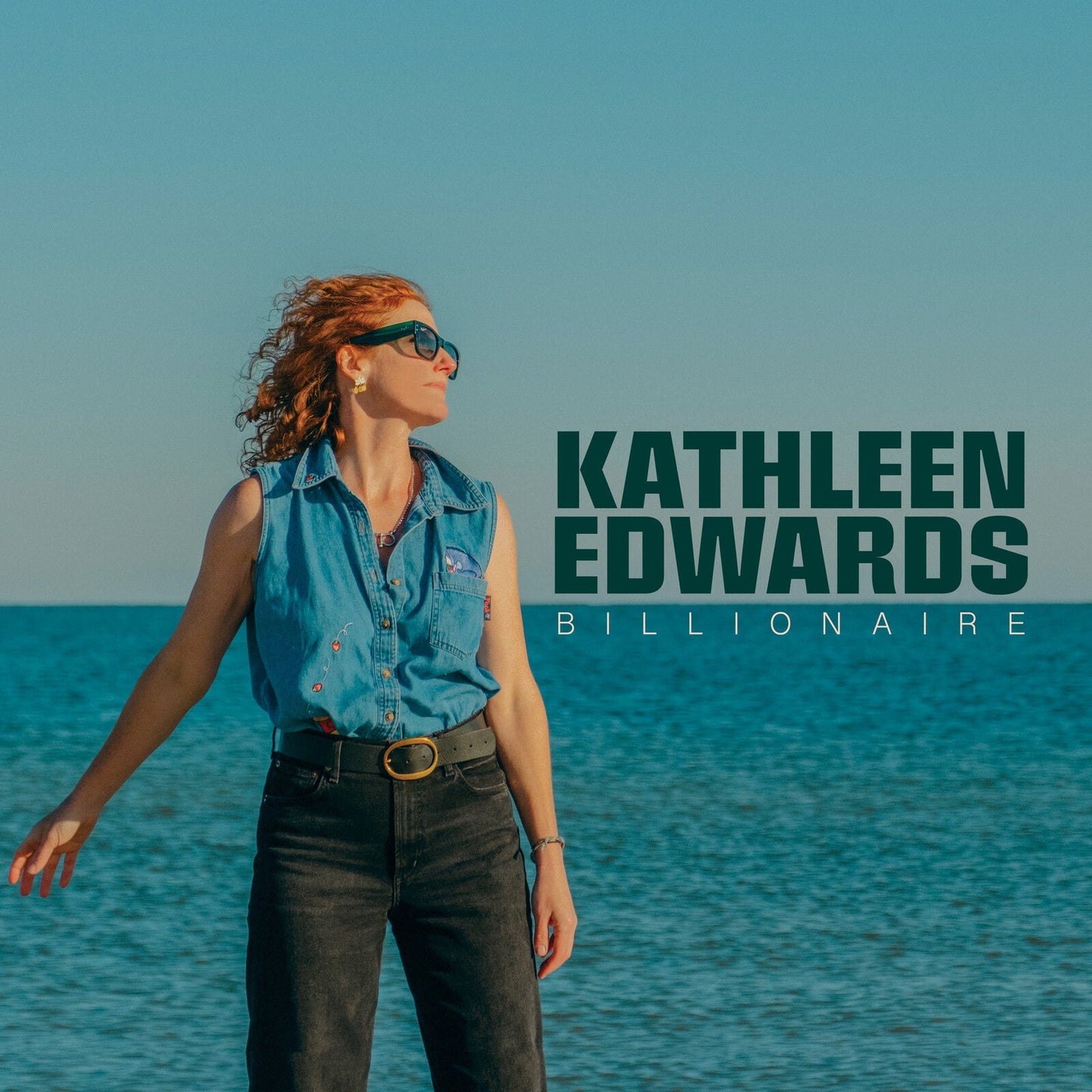 Kathleen Edwards wearing a denim shirt and sunglasses standing before the ocean.