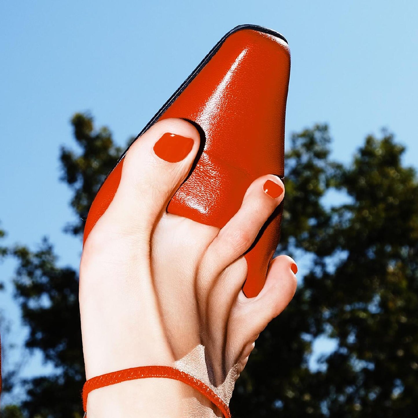 A foot with red nail polish holding a red leather shoe against a sky.