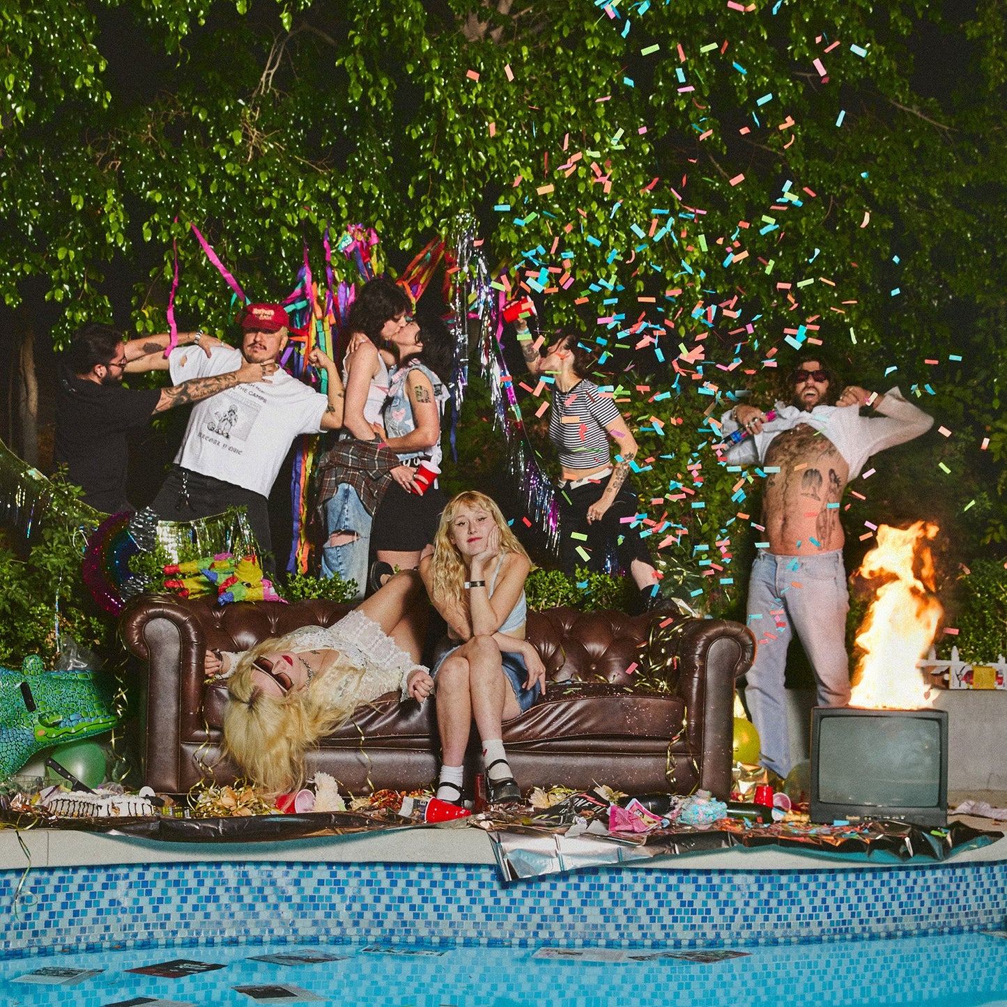A group of people partying by a pool with confetti, a couch, and fire.