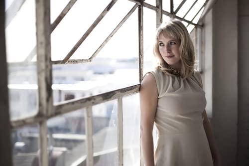 Woman with blonde hair looks thoughtfully from a window.