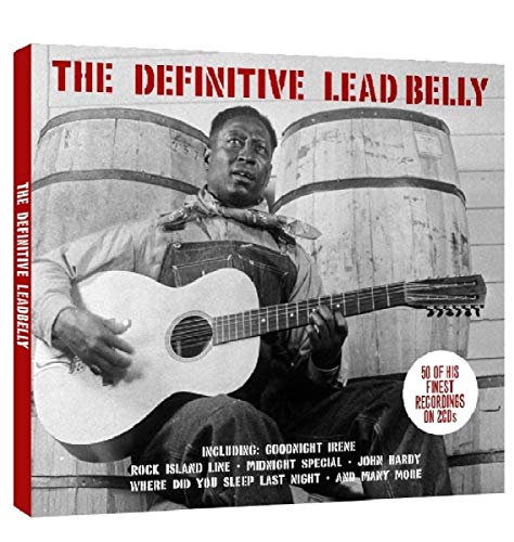 Black and white photo of Lead Belly playing guitar in front of two wooden barrels.
