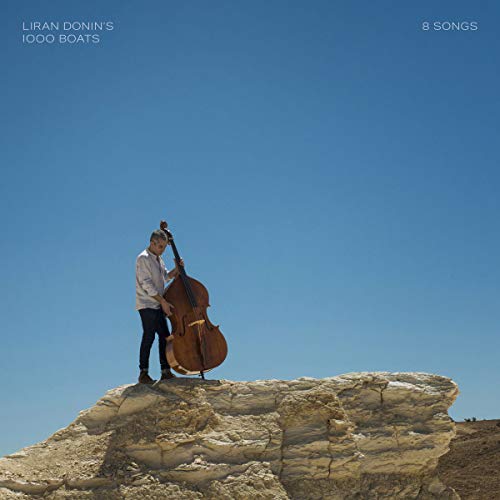 Man stands on rocky outcrop, holding double bass, against clear blue sky, album cover.