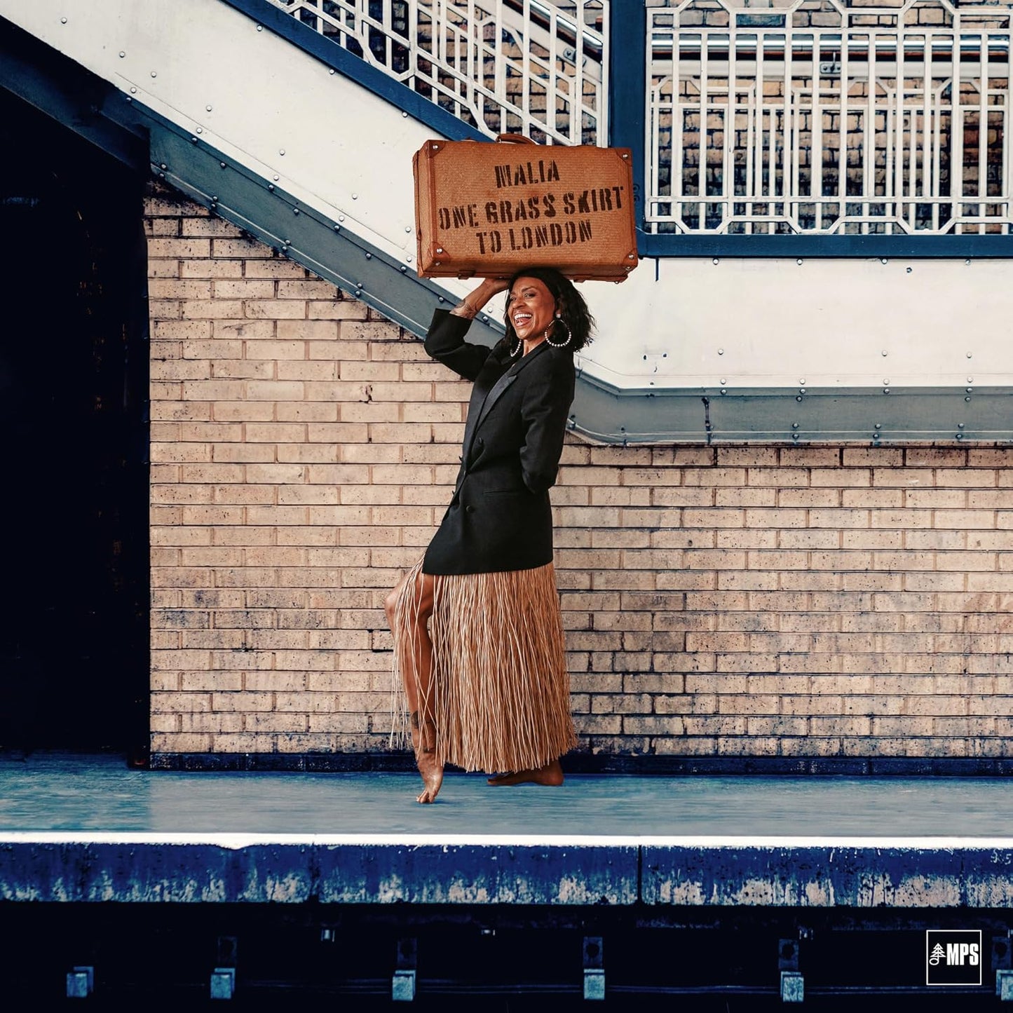 Woman in grass skirt and blazer balancing a suitcase on her head.