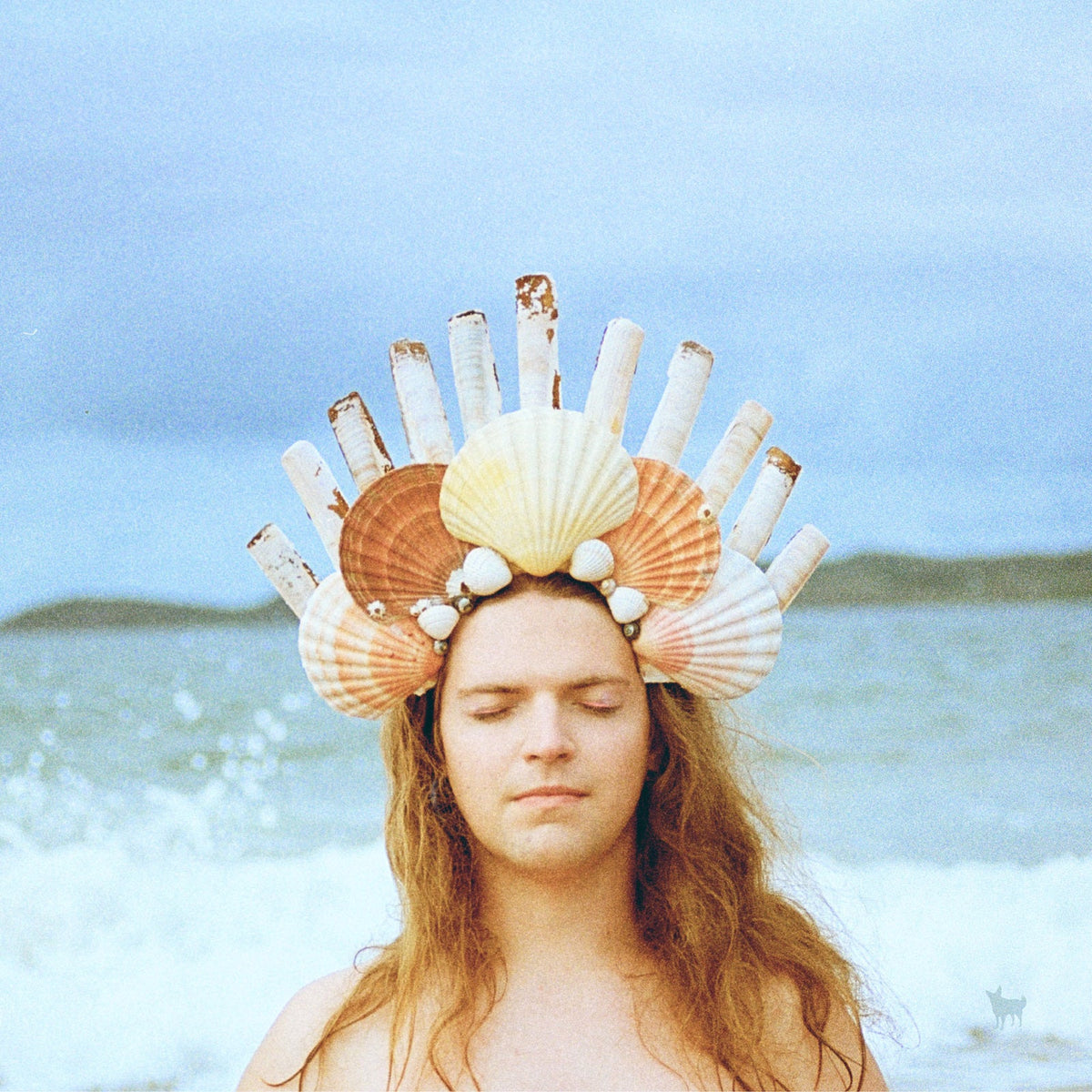 Person with seashell crown, eyes closed, ocean background.
