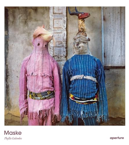 Two figures in textured robes and elaborate horned masks stand outdoors.
