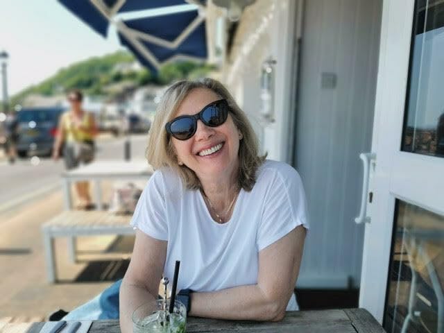 Woman with sunglasses smiling, seated at a table outdoors.