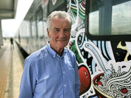 Michael Palin standing on a train platform next to a colorful mural on a train.