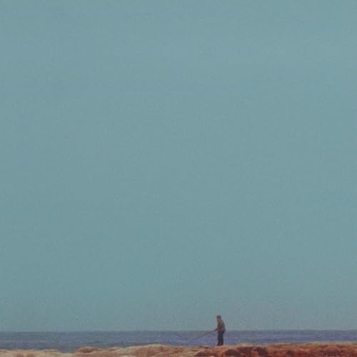 Lone figure fishing on a beach under a pale blue sky.