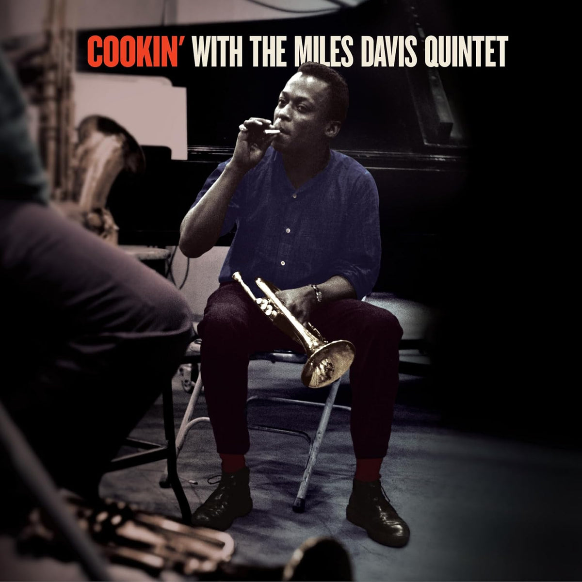 Miles Davis seated, holding a trumpet, with band members in the background.