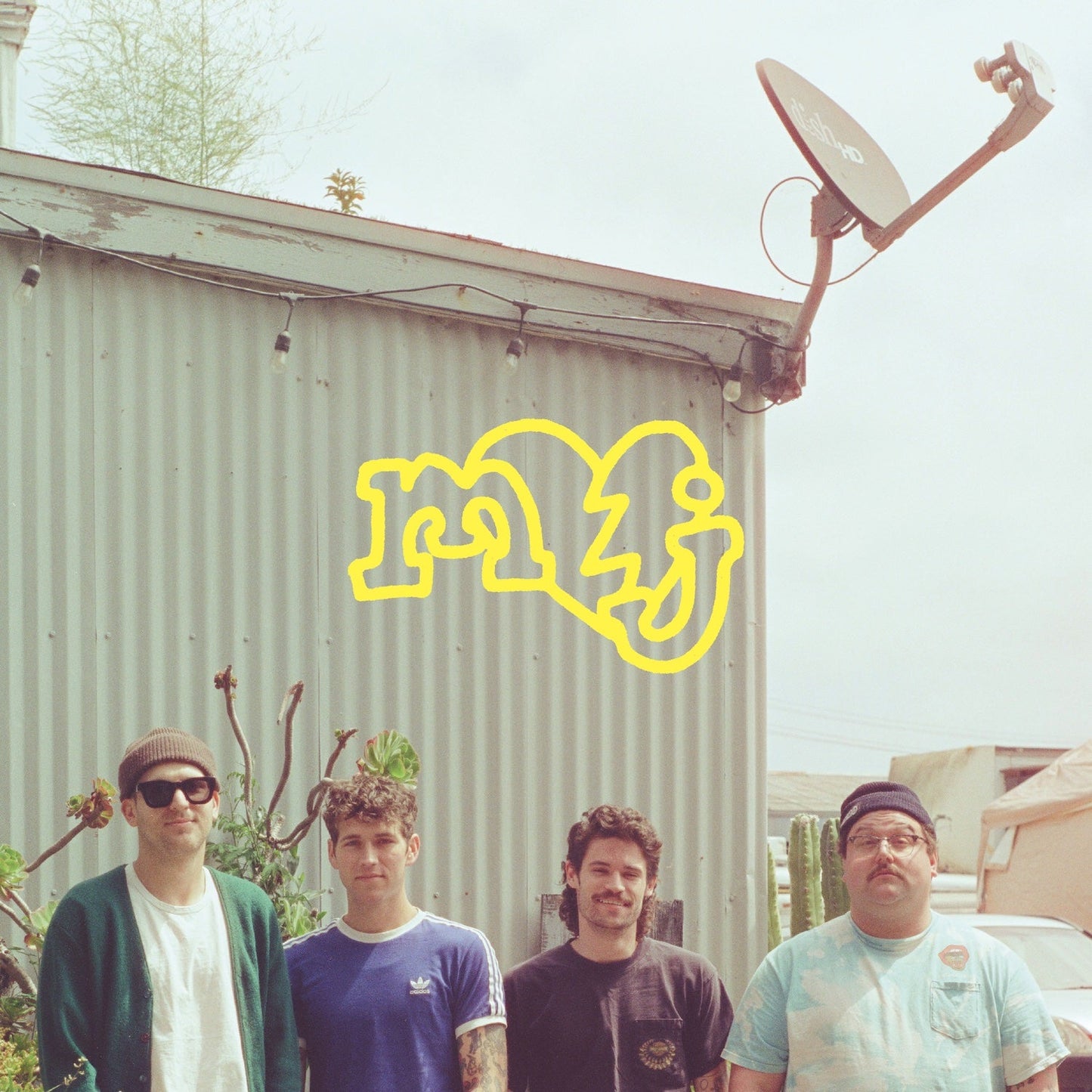 Four men stand before a corrugated metal wall with yellow graffiti.