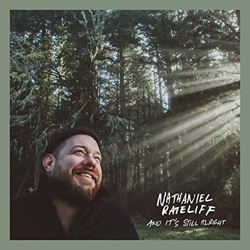 Man in black shirt and hat smiling in forest with sunbeams shining through trees.