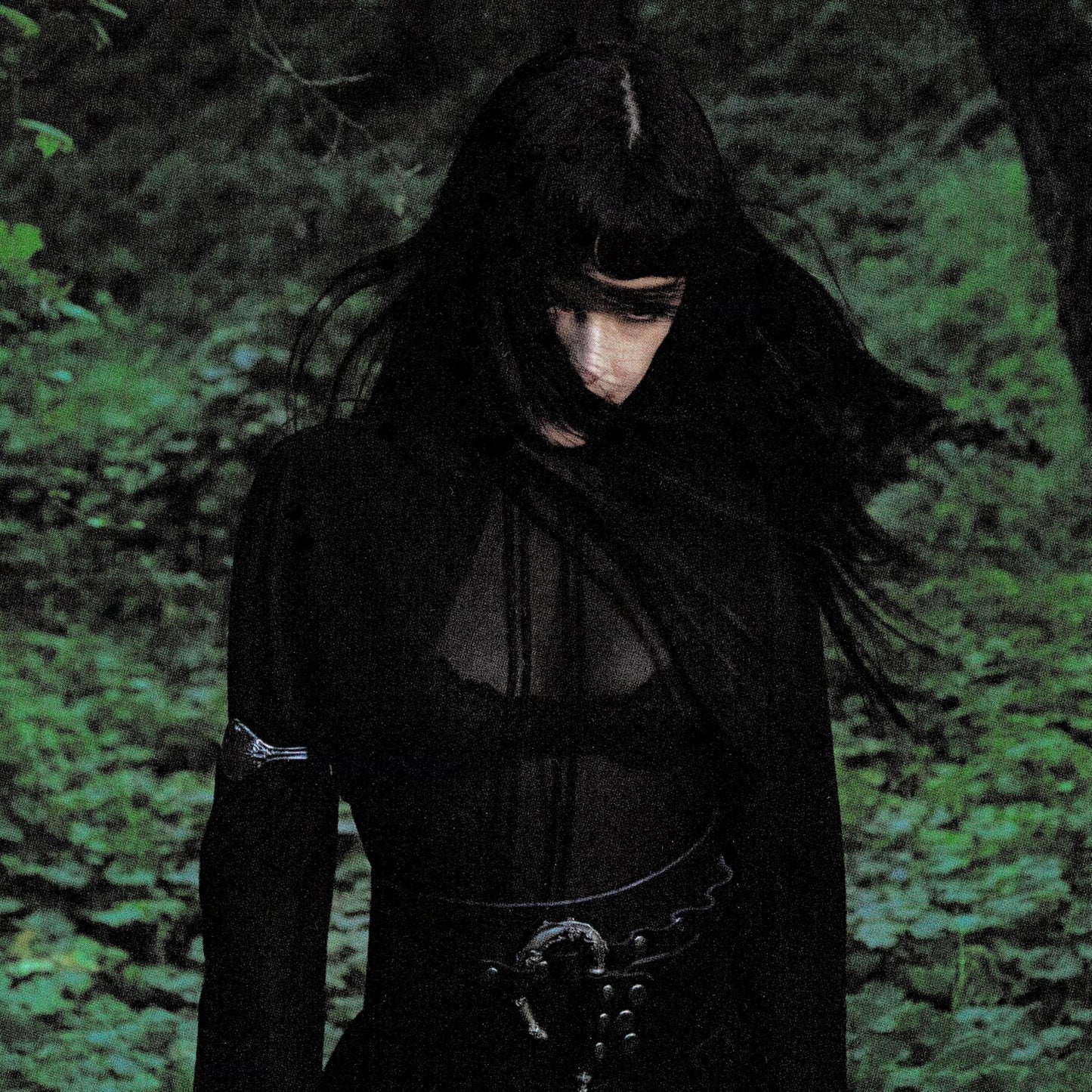 A person with long black hair and dark clothing stands in a green forest.