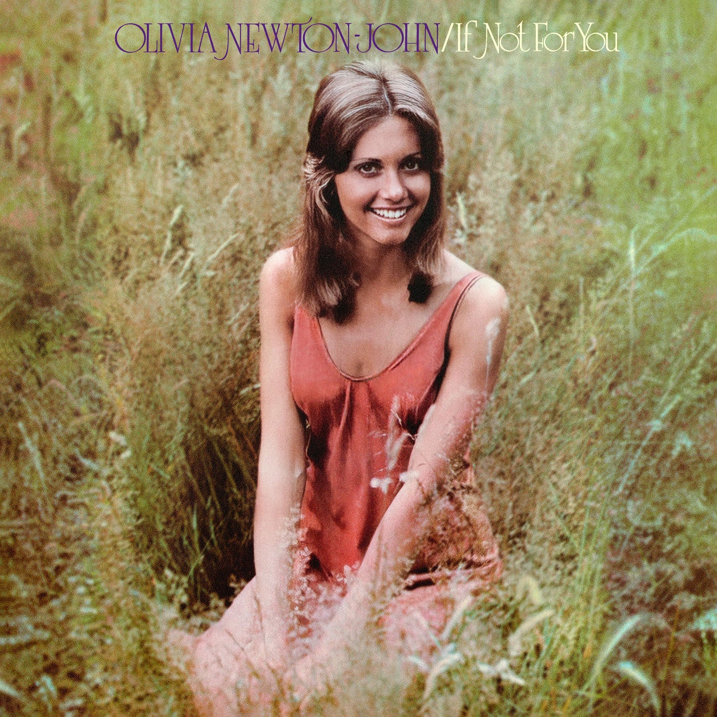 Olivia Newton-John smiling in a red dress while sitting in a grassy field.
