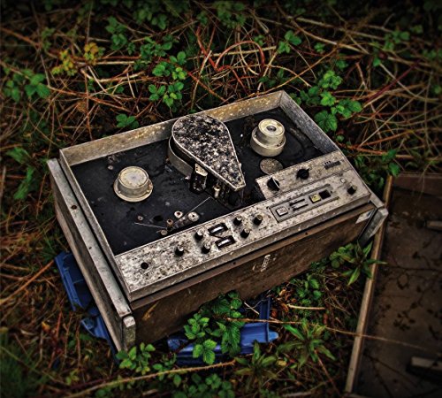 Rusty reel-to-reel tape recorder sits outdoors amongst green foliage.