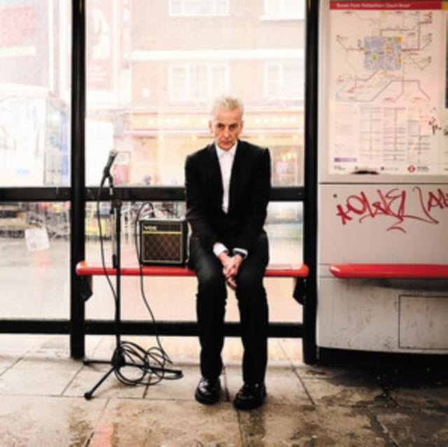 Peter Capaldi in a suit sits at a bus stop with a guitar amp.