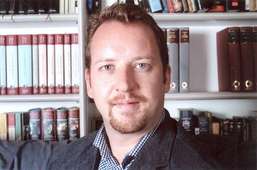 Man with short brown hair and beard, standing in front of bookshelves.