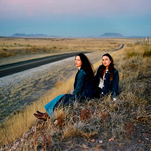 Two women sit side-by-side on a grassy hill overlooking a desert highway.