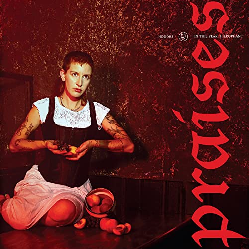 A person with tattoos wears a corset and skirt, holding fruit against a red background.