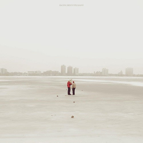 Two figures with antlers stand on a snowy beach, city skyline distant.