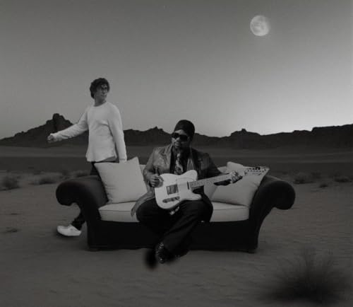 Two musicians with guitars sit on a couch in a desert landscape.