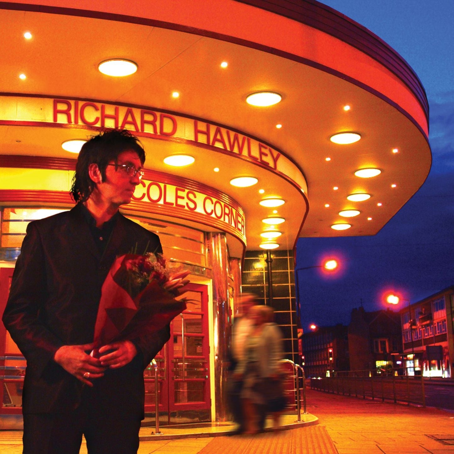 Richard Hawley stands before a brightly lit cinema marquee, holding flowers.