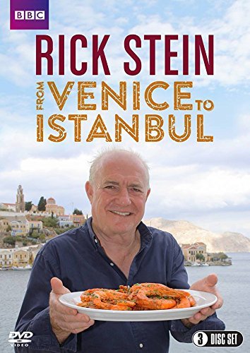 Rick Stein holding a seafood dish, with a coastal town in the background.
