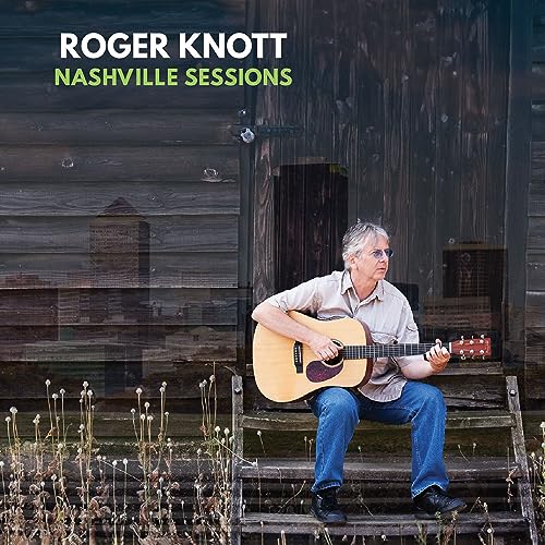 Roger Knott with acoustic guitar, sitting on steps in front of wood siding.
