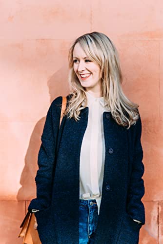 Blonde woman in a dark coat and jeans smiles at the camera.