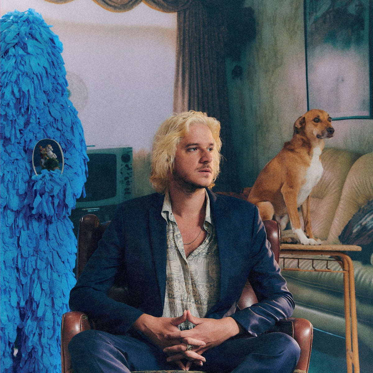 Blond man in suit sits with dog and blue sculpture.