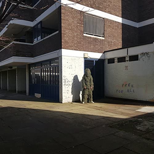 Person in camouflage suit stands before a brick building with graffiti.
