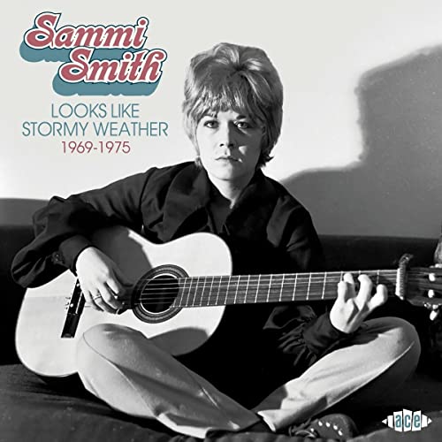 Sammi Smith sits cross-legged, playing an acoustic guitar on a couch.