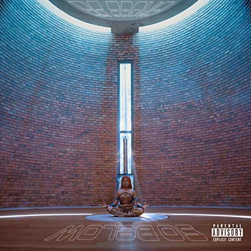 Woman meditates cross-legged on floor in circular room with brick walls and tall window.