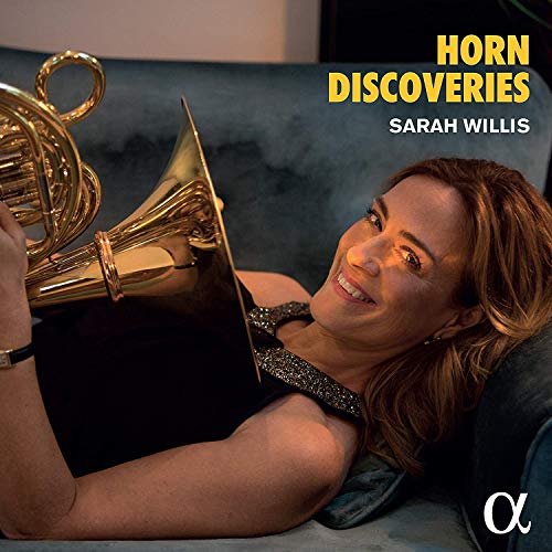 Sarah Willis smiling while holding a French horn on a blue velvet sofa.