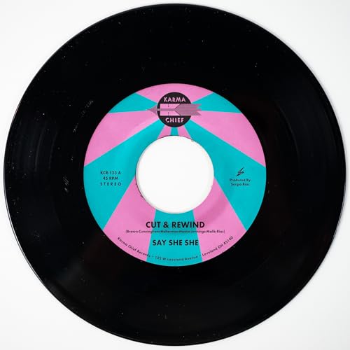 Black vinyl record with a pink and teal center label, reading "Say She She".
