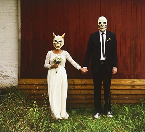 Couple in wedding attire wearing skull and horned masks, holding hands.