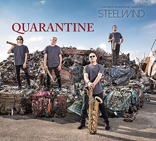 Four musicians with saxophones stand before a large scrap metal pile.