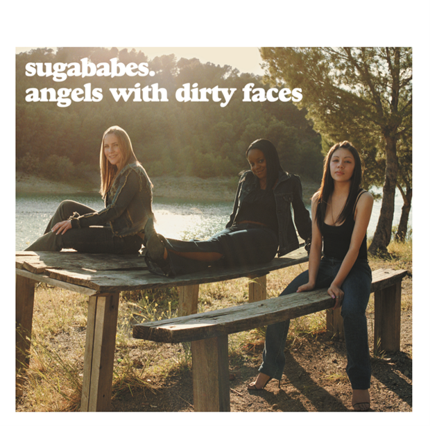 Three women sit on a wooden picnic table outdoors near a lake at sunset.