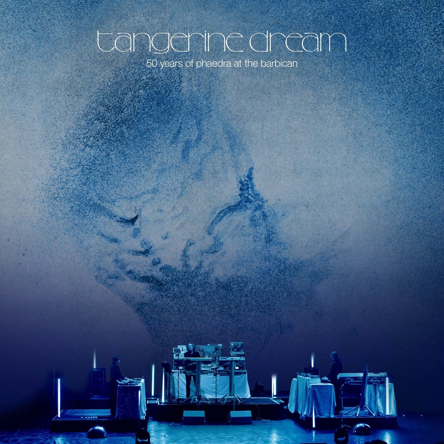 Tangerine Dream performing on stage with a large blue abstract projection in the background.