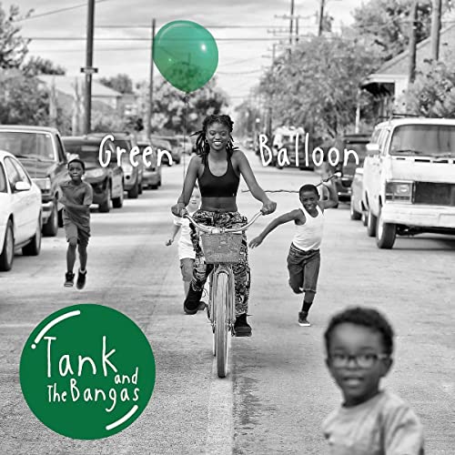 A woman rides a bicycle down a street, followed by children, with a green balloon.