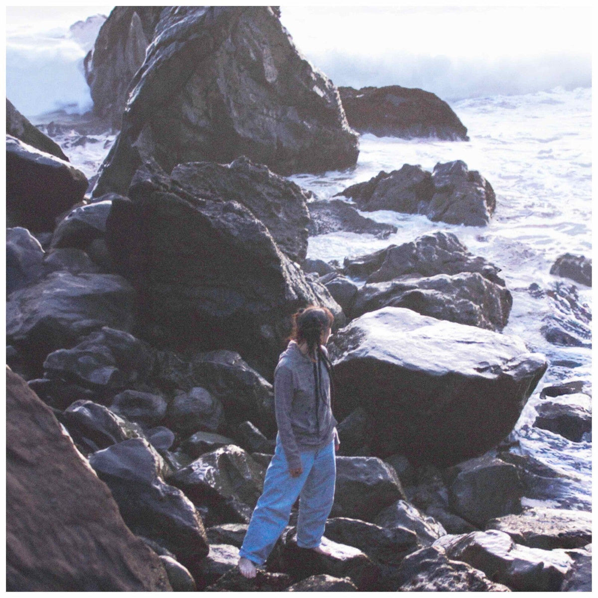 Person standing among rocky coastline with crashing waves in the background.